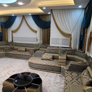 Op de afbeelding: Een luxe woonkamer met een grote hoekbank in tinten grijs en goud. De kamer heeft sierlijke gordijnen in blauw, goud en wit, decoratieve wandpanelen en twee ronde salontafels. Het plafond heeft inbouwverlichting.