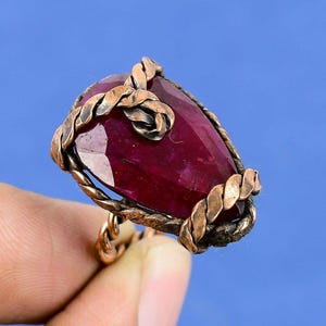 May include: A copper-colored ring with a large, faceted, oval-shaped ruby gemstone. The ruby is a deep red color. The ring's band and setting feature a twisted rope design.