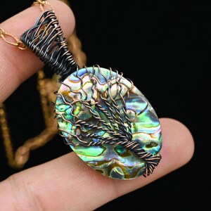 May include: Oval pendant necklace with an abalone shell displaying iridescent green, blue, and gold hues. The shell is encased in a wire-wrapped design, featuring a tree motif. The pendant is suspended from a gold-coloured chain.