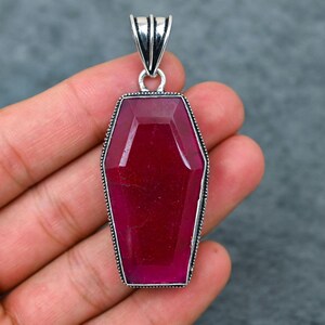 May include: A coffin-shaped pendant with a large, faceted, ruby-red gemstone. The pendant is set in a silver-colored metal with a decorative beaded border and a bail for a chain. The gemstone has a deep, rich color.