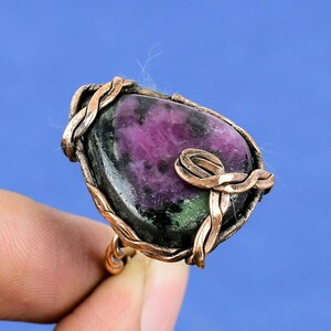 Puede incluir: Anillo artesanal con una gran gema ovalada con tonos morados, verdes y negros. La piedra está engastada en un engaste de alambre de cobre con intrincados detalles trenzados y en bucle, creando un diseño artístico único.