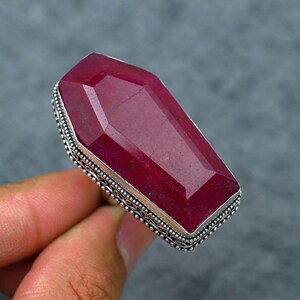 Può includere: Un anello con una grande pietra preziosa di rubino rettangolare. Il rubino è di colore rosso intenso con una superficie sfaccettata. L'anello ha una fascia color argento con un bordo dettagliato e decorato. L'anello è tenuto da una mano su sfondo grigio.