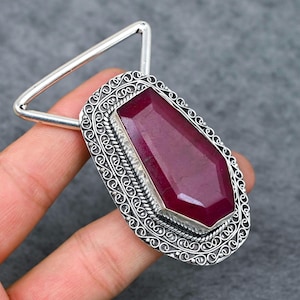 May include: A silver pendant with an oval, ornate frame. The pendant features a large, rectangular, deep red gemstone. The frame has a decorative, swirling pattern. A silver triangle is attached to the top of the pendant.