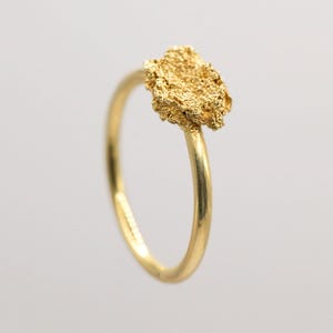 May include: A gold ring with a textured nugget-style design. The ring has a smooth, polished gold band, and the nugget is an irregular shape. The ring is set against a plain white background, which accentuates the gold's colour and texture.