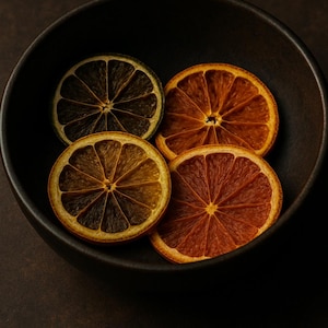 Könnte beinhalten: Eine dunkle, runde Schüssel gefüllt mit vier getrockneten Orangenscheiben. Die Scheiben variieren in der Farbe von hellbraun bis tiefrotbraun, mit sichtbaren radialen Mustern. Die Schüssel steht vor einem dunkelbraunen Hintergrund.