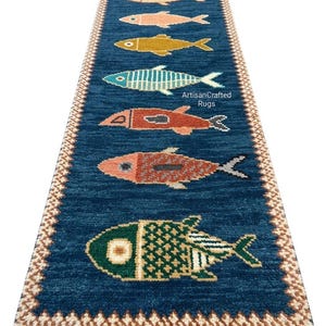 Könnte beinhalten: Ein rechteckiger, blauer Teppich mit einem sich wiederholenden Muster aus bunten Fischen. Die Fische sind in Weiß, Rosa, Gelb, Türkis und Orange gehalten. Der Teppich hat einen Fransenrand und den Text "ArtisanCrafted Rugs".