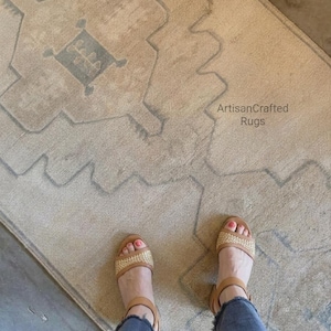 May include: A beige area rug featuring a faded geometric pattern in light grey and a central dark grey rectangular motif. The text ArtisanCrafted Rugs is printed on the surface. The rug has a textured weave and a rectangular shape.