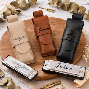 Puede incluir: Tres estuches de cuero para armónica en beige, marrón y negro, cada uno con su armónica a juego. El estuche y la armónica beige están grabados con "Aurora" y un diseño floral. El estuche y la armónica marrón llevan "Jackson". El estuche y la armónica negra llevan "Aaron".