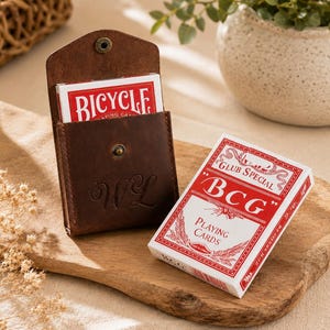May include: A brown leather case with a snap closure and embossed initials WL, containing a deck of red and white Bicycle playing cards. Beside it sits a second deck of red and white BCG Club Special playing cards with white text.