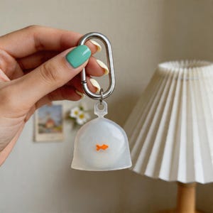 Könnte beinhalten: Ein klarer, fischglasförmiger Schlüsselanhänger mit einem orangefarbenen Fischdesign im Inneren. Der Schlüsselanhänger ist an einem silbernen Metallkarabiner befestigt. Der Hintergrund umfasst einen weißen Lampenschirm und eine Wand mit einem kleinen Bild.