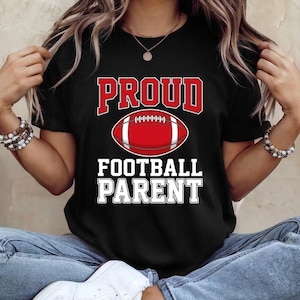 Puede incluir: Camiseta negra con las palabras "PROUD FOOTBALL PARENT" en letras blancas y rojas. Un gráfico de fútbol americano rojo está centrado entre las palabras. La camiseta es de un material suave.