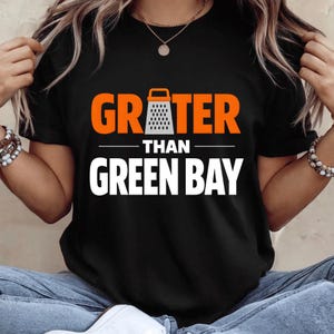 May include: Black t-shirt with the text "GRATER THAN GREEN BAY" in orange and white. The word "GRATER" features a grater graphic. The shirt is a short-sleeved crew neck.
