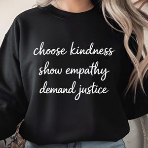 Puede incluir: Sudadera negra con la frase "choose kindness, show empathy, demand justice" en escritura blanca. La sudadera es de color negro liso y tiene cuello redondo. El texto está centrado en la parte delantera.