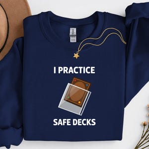 以下が含まれることがあります： 「I PRACTICE SAFE DECKS」の白文字が入ったネイビーブルーのスウェットシャツ。グラフィックは、透明なケースに入った2枚の茶色のカードを示しています。星のペンダントが付いたゴールドのネックレスがスウェットシャツにかけられています。