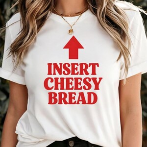 May include: White t-shirt with a red arrow pointing upwards and the text "INSERT CHEESY BREAD" in red, bold letters. The shirt has rolled-up sleeves and is worn by a person.