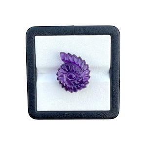 May include: A deep purple amethyst gemstone carved into a spiral ammonite shell shape. The translucent stone features intricate, curved ridges radiating from the center, showcasing a polished finish and vibrant violet hues throughout the piece.