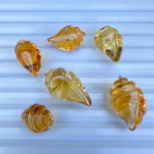 May include: Six amber-coloured glass seashell figurines. The shells have detailed ridges and a glossy finish. The varying shapes and sizes of the shells create a natural, organic aesthetic. The seashells are arranged on a white surface with blue lines.