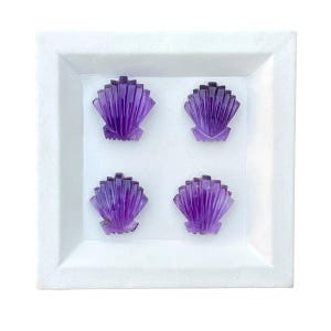 May include: Four carved amethyst gemstone seashell shapes displayed in a white tray. The purple shells have a detailed, fan-like design. The tray is square and has a raised edge, creating a shadow.