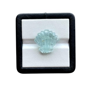 Peut inclure: Une pierre précieuse bleu clair sculptée en forme de coquillage, présentée dans un écrin noir et blanc. La pierre précieuse est translucide, avec des lignes et des arêtes visibles. La boîte est carrée avec un extérieur noir et un intérieur blanc.