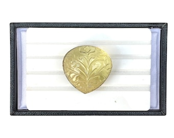 May include: A heart-shaped, yellow-toned brooch with an engraved floral design, presented in a black display box with a white interior. The brooch's intricate details are highlighted against the contrasting background.