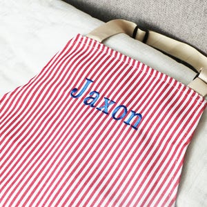 May include: Red and white striped apron with the name "Jaxon" embroidered in blue thread. The apron has a beige strap and is made of a lightweight fabric. The design is simple and classic, suitable for cooking or crafting.
