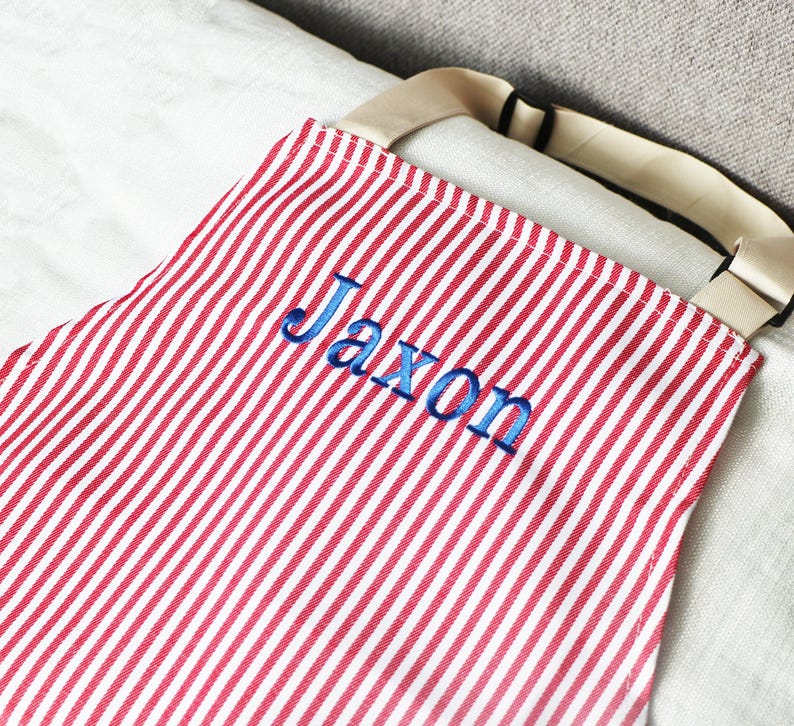 May include: Red and white striped apron with the name "Jaxon" embroidered in blue thread. The apron has a beige strap and is made of a lightweight fabric. The design is simple and classic, suitable for cooking or crafting.