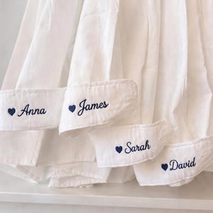 May include: White dress shirts with personalised cuffs. Each cuff features a name embroidered in navy blue with a navy blue heart symbol. The names are Anna, James, Sarah, and David.