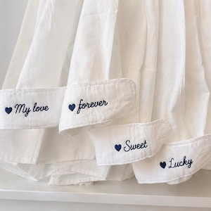 Puede incluir: Primer plano de pu&ntilde;os de camisa blancos con bordados en azul marino. Los pu&ntilde;os muestran las palabras "My love", "forever", "Sweet" y "Lucky". La camisa es de tela blanca suave.