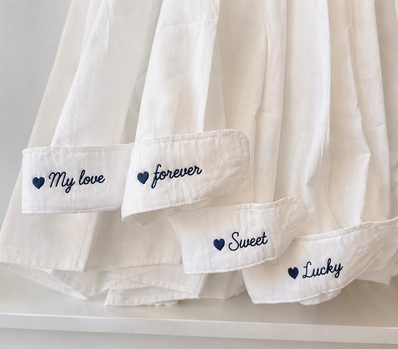 Puede incluir: Primer plano de pu&ntilde;os de camisa blancos con bordados en azul marino. Los pu&ntilde;os muestran las palabras "My love", "forever", "Sweet" y "Lucky". La camisa es de tela blanca suave.