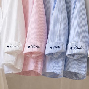 Puede incluir: Camisas de pijama a rayas blancas, rosas y azules colgadas en una percha. Cada pu&ntilde;o est&aacute; bordado con un nombre o t&iacute;tulo: Emma, Olivia, mam&aacute;, Novia y Sophie. Una botella y una copa de champ&aacute;n est&aacute;n sobre una mesa.