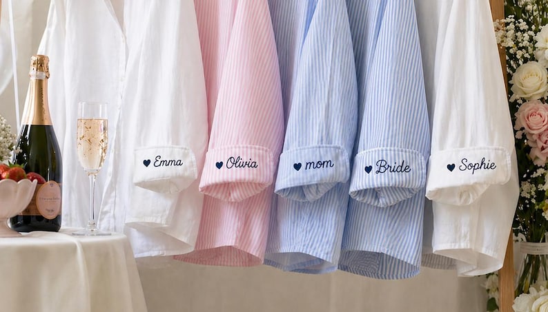 Puede incluir: Camisas de pijama a rayas blancas, rosas y azules colgadas en una percha. Cada pu&ntilde;o est&aacute; bordado con un nombre o t&iacute;tulo: Emma, Olivia, mam&aacute;, Novia y Sophie. Una botella y una copa de champ&aacute;n est&aacute;n sobre una mesa.