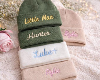 May include: Stack of personalized beanies in olive green, white, and tan. The beanies are embroidered with names and phrases in various colors. A pink rose and sprigs of white flowers are in the background.