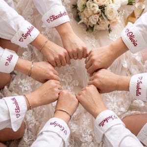 Puede incluir: Camisas blancas abotonadas con nombres personalizados bordados en rojo en los pu&ntilde;os. Los nombres incluyen Mia, Elizabeth, Sophia, Olivia, Isabella y Evelyn. Las camisas est&aacute;n dispuestas en c&iacute;rculo.