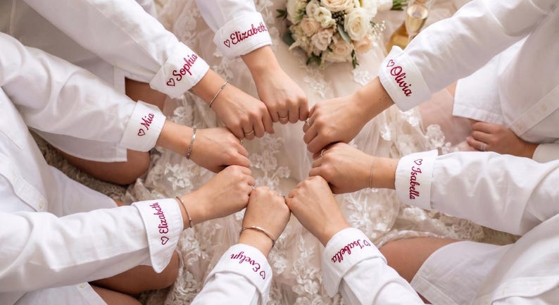 Puede incluir: Camisas blancas abotonadas con nombres personalizados bordados en rojo en los pu&ntilde;os. Los nombres incluyen Mia, Elizabeth, Sophia, Olivia, Isabella y Evelyn. Las camisas est&aacute;n dispuestas en c&iacute;rculo.