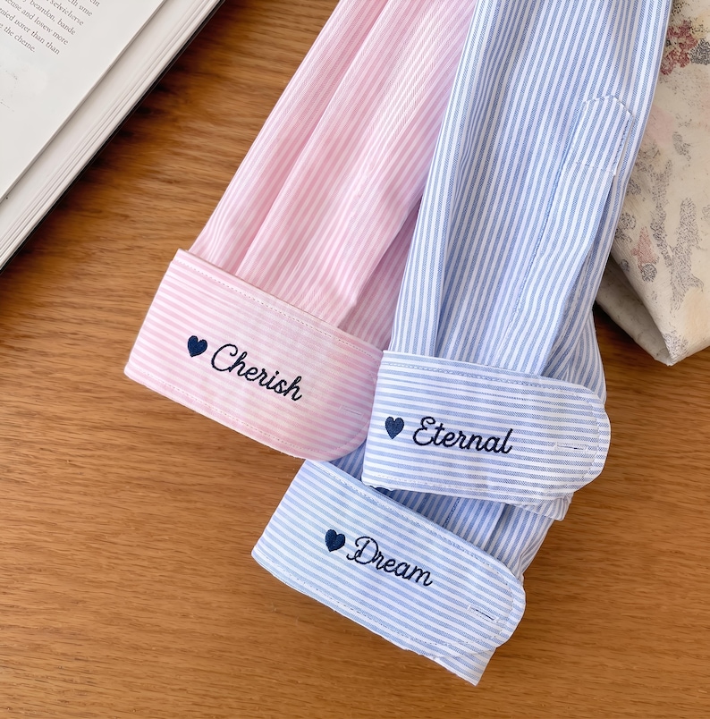 Puede incluir: Primer plano de tres mangas de camisa a rayas. Una es rosa con la palabra "Cherish" bordada en el pu&ntilde;o. Las otras dos son azules y blancas, con "Eternal" y "Dream" bordadas en los pu&ntilde;os. Cada pu&ntilde;o tiene un peque&ntilde;o coraz&oacute;n.