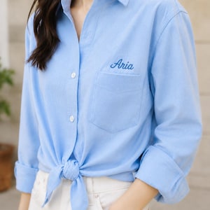 Puede incluir: Camisa abotonada azul claro con el nombre "Aria" bordado en el bolsillo. La camisa est&aacute; atada a la cintura y tiene las mangas enrolladas. El modelo lleva pantalones blancos.