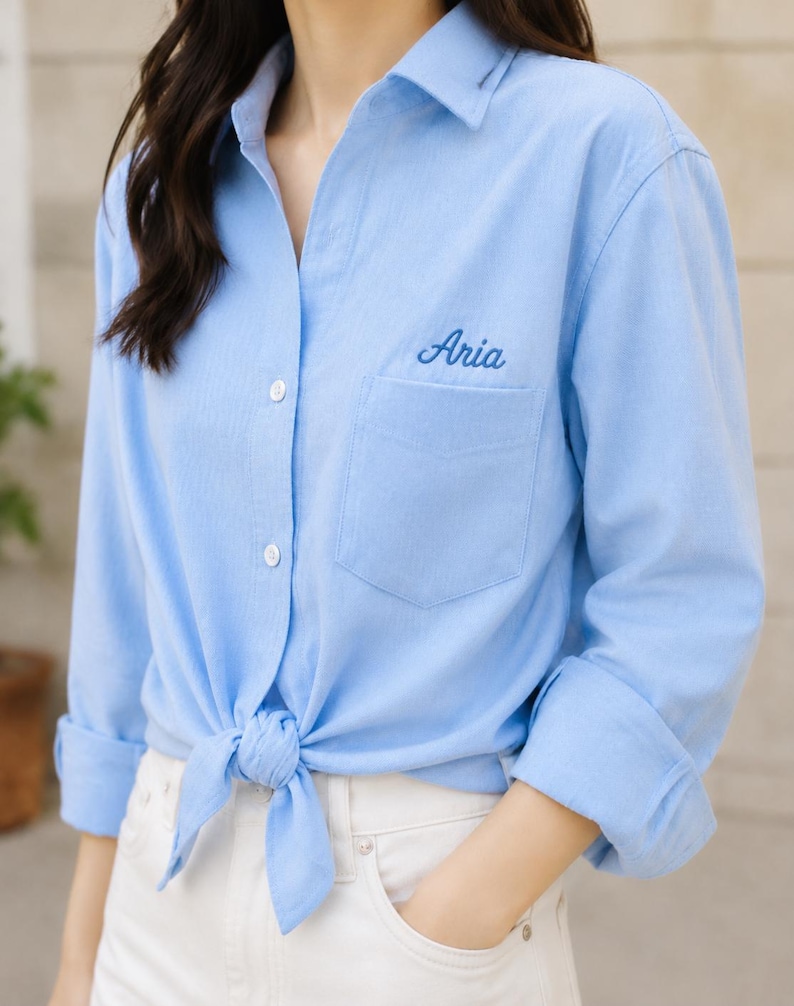 Puede incluir: Camisa abotonada azul claro con el nombre "Aria" bordado en el bolsillo. La camisa est&aacute; atada a la cintura y tiene las mangas enrolladas. El modelo lleva pantalones blancos.