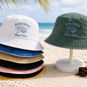 Op de afbeelding: Een stapel kleurrijke vissershoeden in wit, blauw, beige, zwart, roze en geel, en een groene hoed op een standaard. Elke hoed heeft de tekst "Bahamas Beach Club" in blauw. Zonnebrillen liggen op het zand.