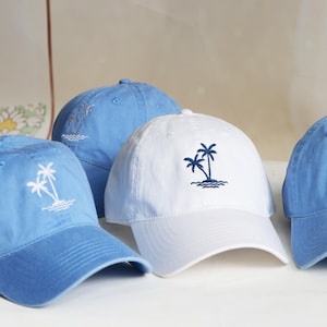 Op de afbeelding: Een collectie van vier baseballpetten in lichtblauwe en witte tinten. Elke pet is voorzien van een geborduurd ontwerp van twee palmbomen op een klein eiland met golvende waterlijnen. Het borduurwerk is wit op de blauwe petten en donkerblauw op de witte.
