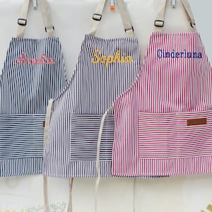 May include: Three striped aprons with personalized names embroidered on the front. The aprons are navy blue, purple, and red with white stripes. Each apron has a front pocket and adjustable straps.