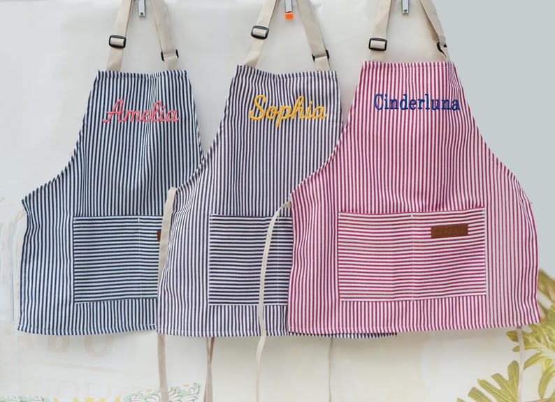 May include: Three striped aprons with personalized names embroidered on the front. The aprons are navy blue, purple, and red with white stripes. Each apron has a front pocket and adjustable straps.