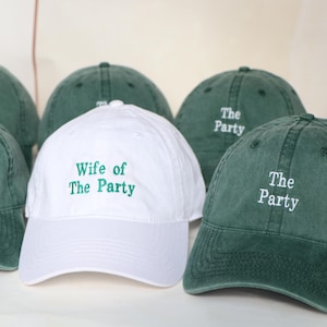 May include: A selection of green baseball caps with white embroidered text reading "The Party." One white cap is embroidered with "Wife of The Party." The caps are made of a soft fabric and have a curved peak.