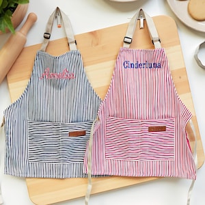 May include: Two striped aprons, one navy and white, the other red and white, with embroidered names "Amelia" and "Cinderluna". Each apron has a front pocket and adjustable straps. They are displayed on a wooden cutting board, with baking supplies in the background.