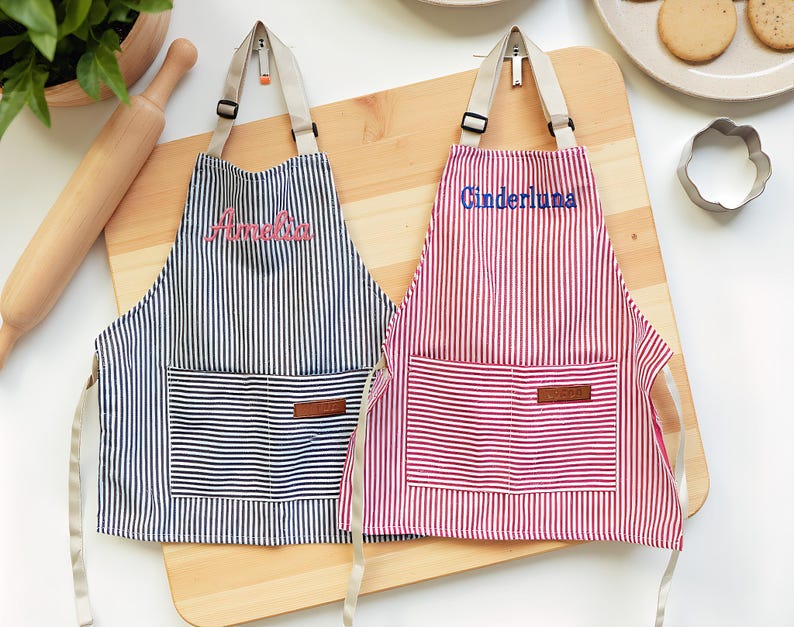 May include: Two striped aprons, one navy and white, the other red and white, with embroidered names "Amelia" and "Cinderluna". Each apron has a front pocket and adjustable straps. They are displayed on a wooden cutting board, with baking supplies in the background.
