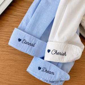 Puede incluir: Primer plano de tres pu&ntilde;os de camisa. Dos son de color azul claro, uno es blanco. Cada pu&ntilde;o tiene bordado el texto: "Eternal", "Cherish" y "Dream", con un s&iacute;mbolo de coraz&oacute;n delante de cada palabra. Los pu&ntilde;os est&aacute;n sobre una superficie de madera.