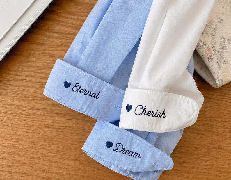 Puede incluir: Primer plano de tres pu&ntilde;os de camisa. Dos son de color azul claro, uno es blanco. Cada pu&ntilde;o tiene bordado el texto: "Eternal", "Cherish" y "Dream", con un s&iacute;mbolo de coraz&oacute;n delante de cada palabra. Los pu&ntilde;os est&aacute;n sobre una superficie de madera.