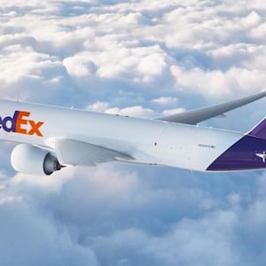 May include: A white FedEx cargo plane flies through a cloudy sky. The plane has the FedEx logo in purple and orange on the fuselage and tail. The tail is purple with the FedEx logo. The plane's wings are silver.