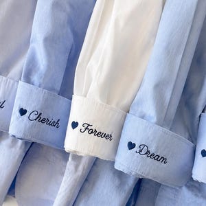 Puede incluir: Primer plano de varios pu&ntilde;os de camisa azul claro y blancos. Cada pu&ntilde;o est&aacute; bordado con una palabra y un coraz&oacute;n: "Eternal", "Cherish", "Forever", "Dream" y "Always". Las camisas est&aacute;n cuidadosamente dispuestas.