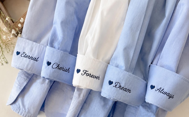 Puede incluir: Primer plano de varios pu&ntilde;os de camisa azul claro y blancos. Cada pu&ntilde;o est&aacute; bordado con una palabra y un coraz&oacute;n: "Eternal", "Cherish", "Forever", "Dream" y "Always". Las camisas est&aacute;n cuidadosamente dispuestas.