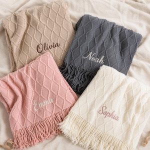 May include: Four knitted blankets in beige, dark grey, dusty pink, and cream, each featuring a diamond pattern, fringe edges, and embroidered names: Olivia, Noah, Emma, and Sophia. The blankets are arranged in a group on a soft fabric surface.